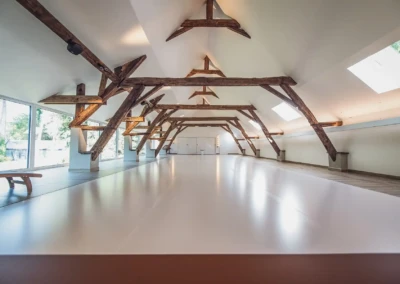 Grande salle avec poutres en bois apparentes, plafond blanc, fenêtres panoramiques et sol en bois clair, éclairée par des puits de lumière.