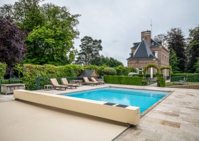 Piscine entourée de transats beige et d'une terrasse en pierre, avec un château en briques rouges à l'arrière-plan et des arbres verdoyants.