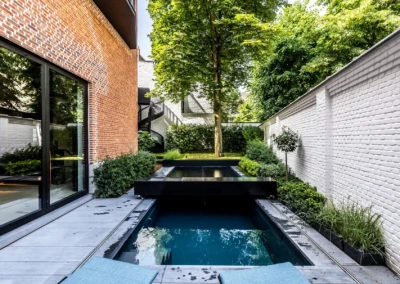Une piscine rectangulaire entourée de murs en briques rouges et blanches, avec des plantes vertes et un arbre au fond.