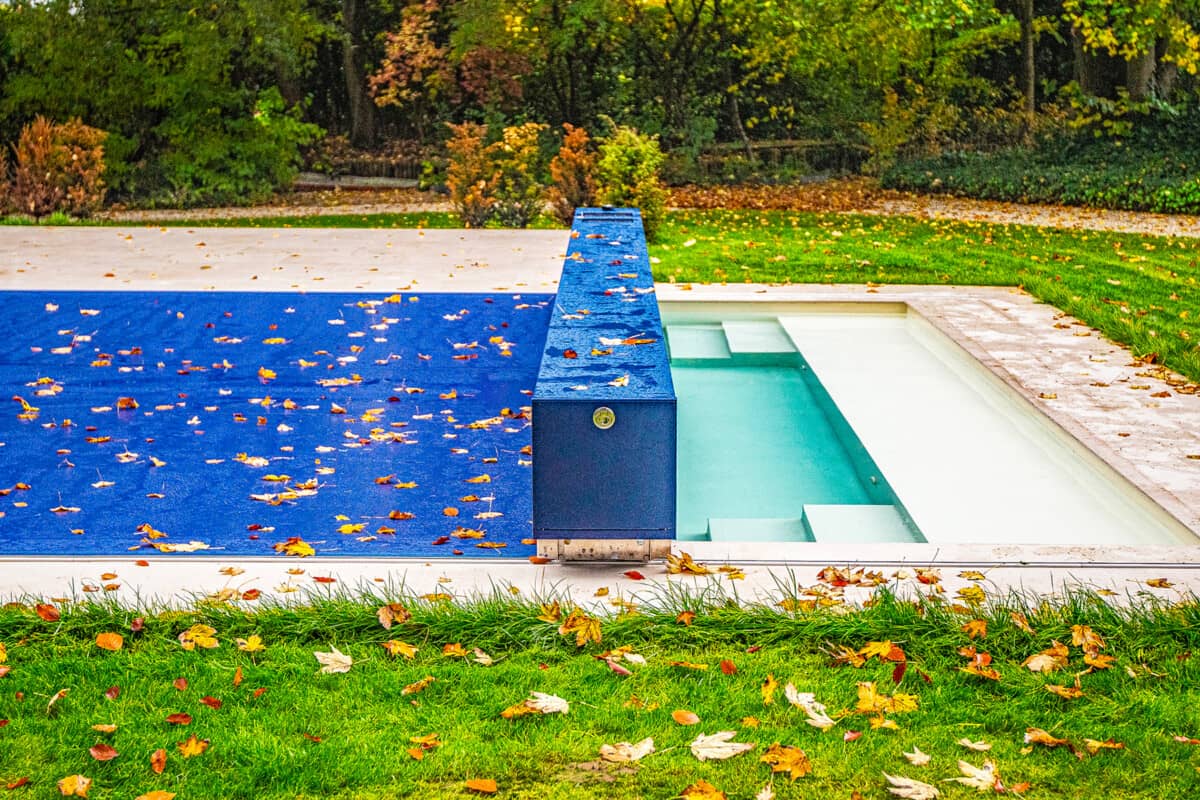 Couverture de piscine bleue avec feuilles mortes, entourée de pelouse verte et arbres en arrière-plan.