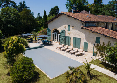 Piscine rectangulaire couverte par une Coverseal grise, entourée de transats et d’une maison en pierre au style traditionnel avec volets verts.